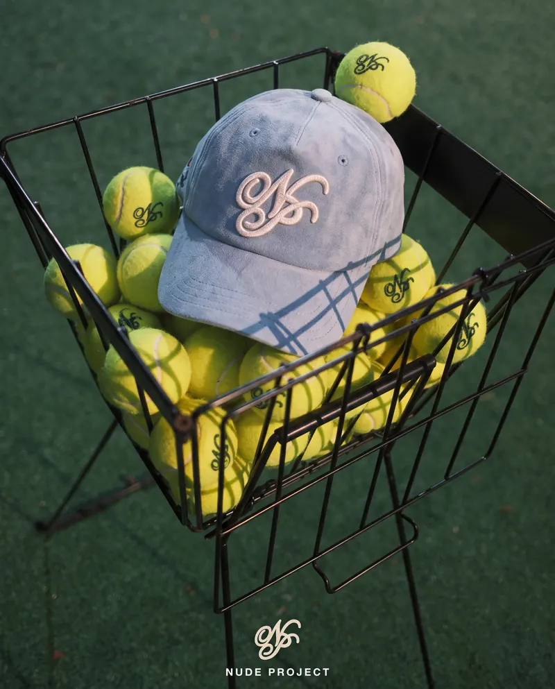 Light blue Nude Project cap resting on tennis balls in a wire ball basket on a green court