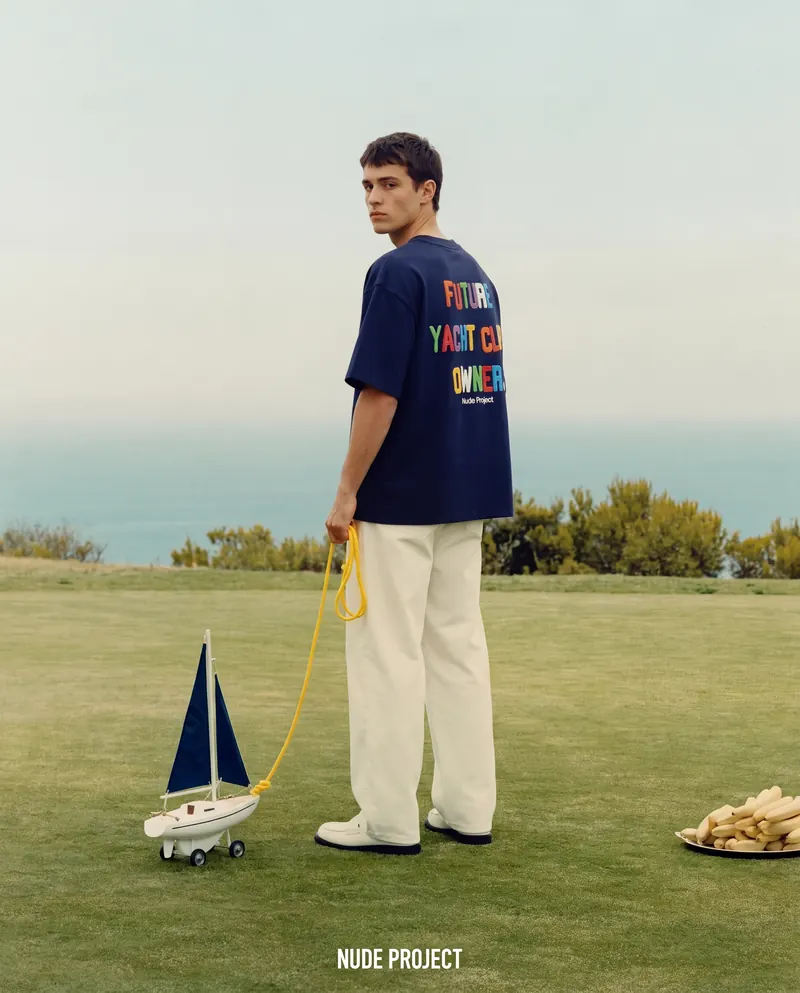 Model in a navy Nude Project tee on a clifftop lawn, holding a toy sailboat on a yellow line